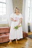 Woman in a white dress holding flowers in a room with large windows and a pink couch.