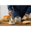 Person spreading butter on bread with a knife, surrounded by bread and small containers on a wooden board.