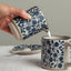Person pouring milk from a blue and white patterned ceramic pitcher into another similar mug on a neutral background.