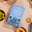 A package of Pea Green Boat Bakery Cheese Sablés Original is shown in a styled flat-lay on a pink hexagonal tile surface, surrounded by green olives, a cracker, and a cheese dip bowl.