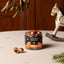 A clear glass jar of Lakrids 'Bulow' Classic food product sits on a light beige tabletop, with two copper-colored round candies placed beside it. The background features a warm wooden wall and a decorative white rocking horse, creating a cozy holiday atmosphere.
