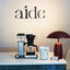 Coffee-making equipment and 'aide' branded items on a counter with 'aide' logo in the background.