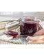 A clear glass clamp-lid jar filled with dark red jam, set on a reflective silver tray with a spoonful of jam and a folded pale pink cloth nearby.