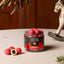 A clear jar of Lakríds by Bølow Raspberry gummy candies sits on a light tabletop with a few candies placed beside it. The jar has a black label with red raspberry graphics, and the background includes a wooden door and a decorative rocking horse.