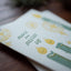 A close-up of a printed card or paper with the phrase “MAKE THE YULETIDE GAY” in green lettering, decorated with yellow and green holiday illustrations, resting on a dark wooden surface.