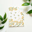 A white rectangular card or notebook cover with a botanical illustration pattern of yellow-orange flowers and green stems, plus the text 'THINKING OF YOU' in the top right, styled on a white surface with green leaves around it.