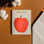 A square white greeting card with a cute red apple illustration and the words 'Thank you' in green, styled on a warm brown surface with cotton bolls and a white envelope nearby.