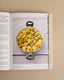 An open cookbook spread showing a recipe page with a photo of stuffed green beans in a pan, photographed against a warm beige background.