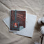 A square greeting card with a white envelope, placed on a beige fabric surface with cotton bolls at the side. The card features a dark brown background with a teal fireplace illustration and orange script reading 'Warm wishes'.