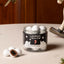 A clear glass jar of Lakrlds by Bülow Frozen Mint chocolate-covered candies sits on a light beige tabletop with a few loose mint candies in front. The jar has a black label with white text and graphics, and the background shows a warm wooden panel and part of a rocking horse.