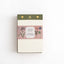 A square paper notebook or journal with an off-white cover, a dusty pink floral paper band reading 'JOTTER BLOCK FESTIVAL OF FLOWERS,' and an olive green top strip with small gold details, photographed on a clean white background.