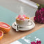 Pink axolotl-shaped tea infuser in a cup of tea on a wooden surface with a flower and fruit.