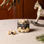 A jar of Lakrids by Bulow Cherry Sour chocolates sits on a light tabletop with a few loose chocolates beside it. The scene includes a wooden rocking horse and hanging greenery in the background, making this a styled product shot rather than a plain studio packshot.