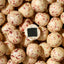 A close-up pile of round cream-colored chocolate truffles speckled with red berry pieces, with one truffle cut open to show a dark chocolate center.