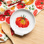 A white ceramic bowl with a painted red tomato on the front, styled on a light wood surface with a matching tomato-print cloth and a wooden spoon.
